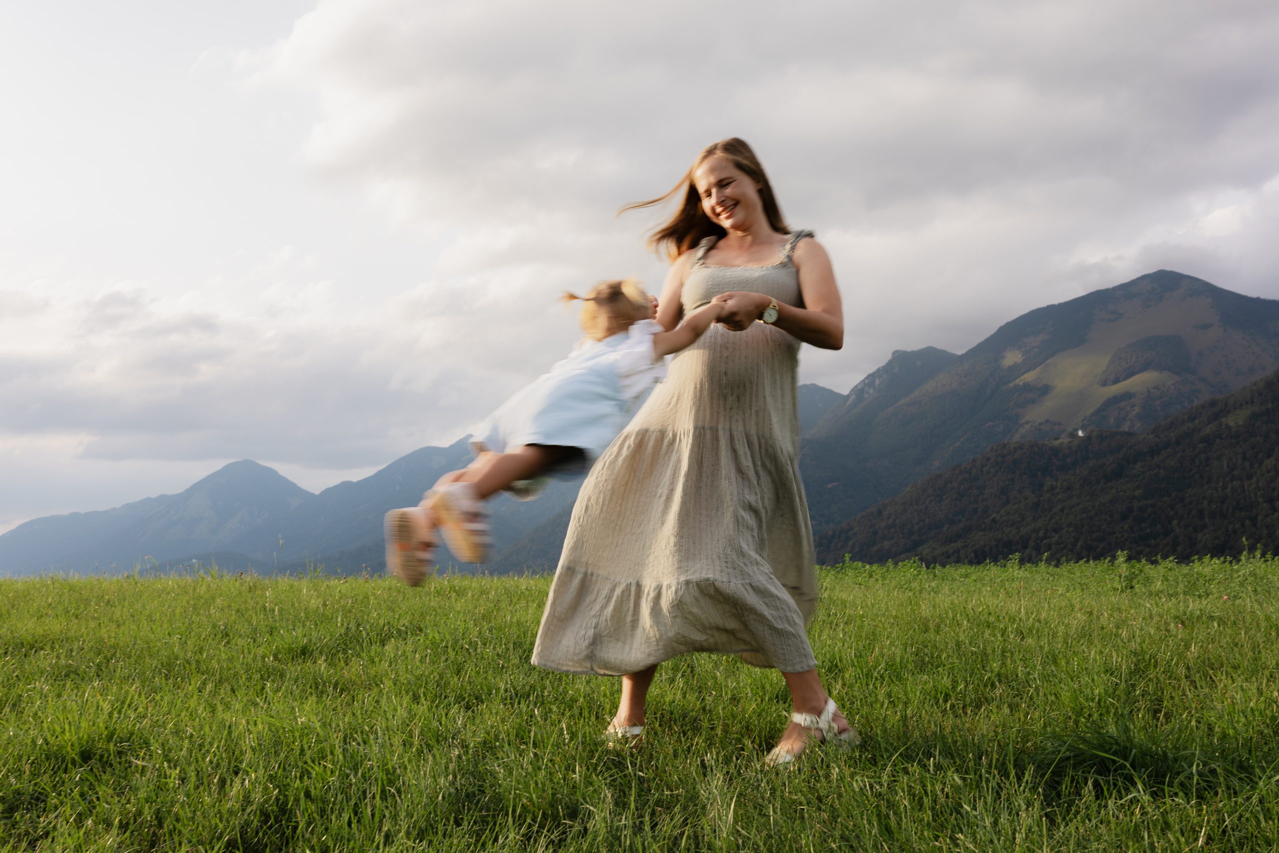Mother spinning with her daughter on a meadow with mountains in the background, family photography Slovenia