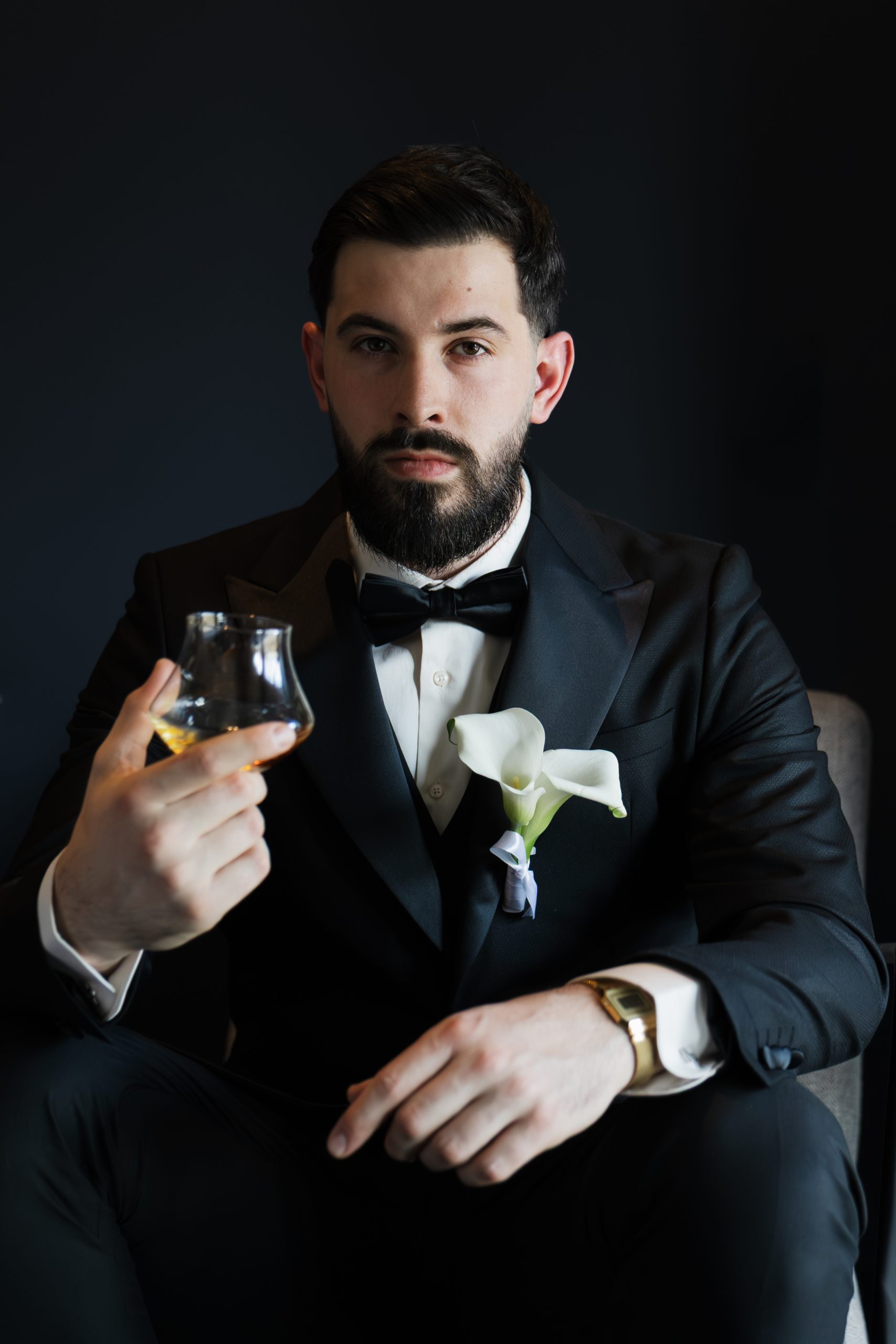 Groom in a black tuxedo holding a glass of whiskey, elegant wedding portrait with dark background