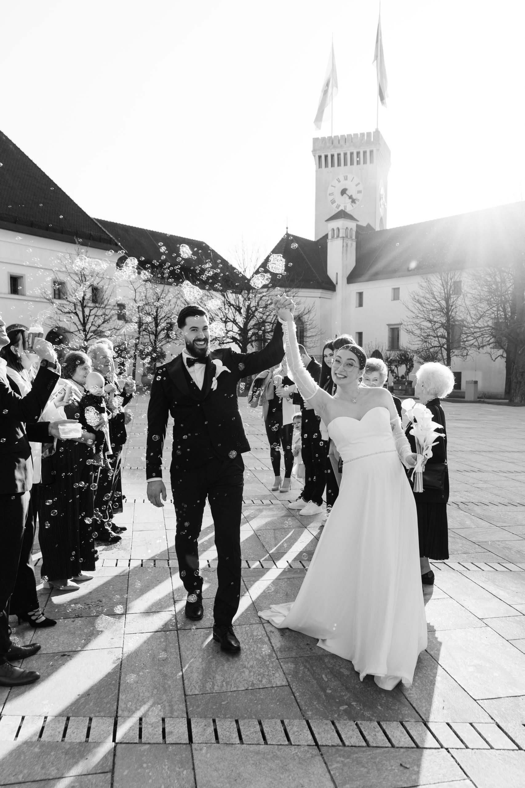Mladoporočenca hodita iz poročnega obreda pred Ljubljanskim gradom, svatje ju obmetavajo z milnimi mehurčki.